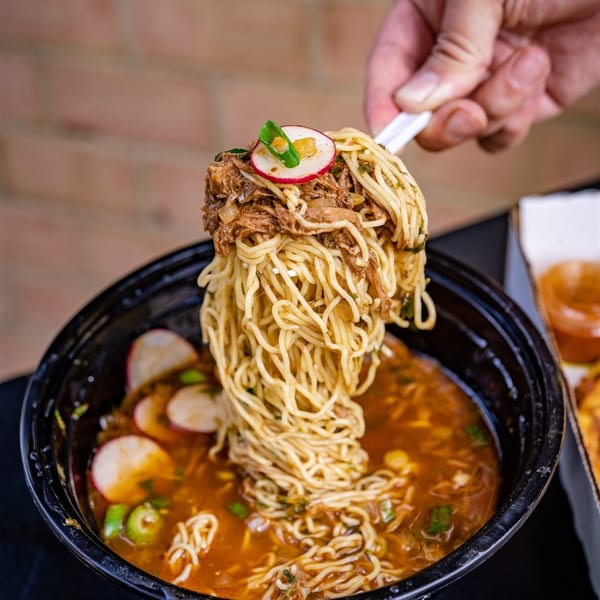 A hand lifts a forkful of Big Papi's birria ramen, topped with shredded meat and a radish slice.