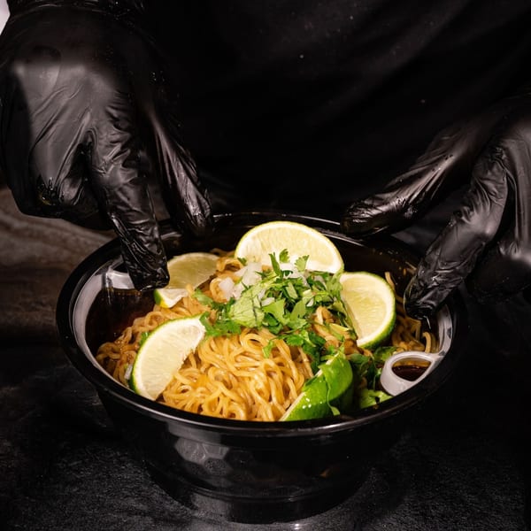 Hands in black gloves holding a bowl of ramen noodles with lime, cilantro, and onions.