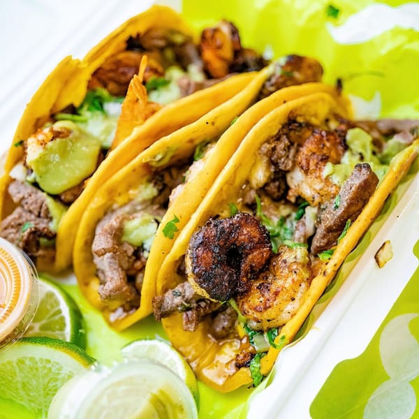 Three Big Papi's tacos with steak, shrimp, guacamole, and lime wedges in a takeout container.