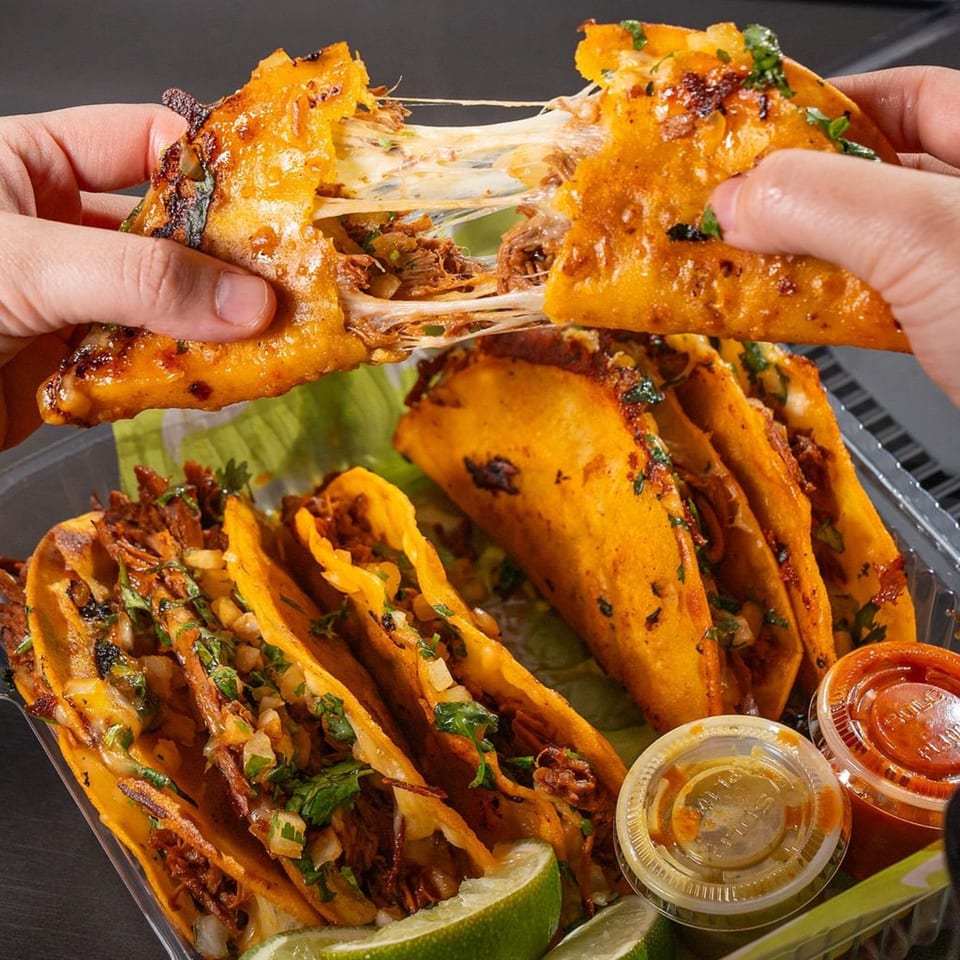 Hands pulling apart a Big Papi's birria taco, revealing melted cheese and shredded beef, with more tacos and lime.