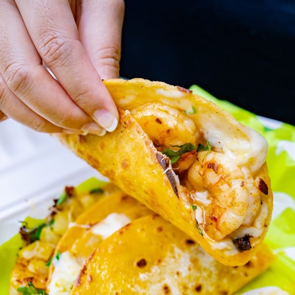 A hand holds a Big Papi's shrimp taco with melted cheese and cilantro over more tacos.