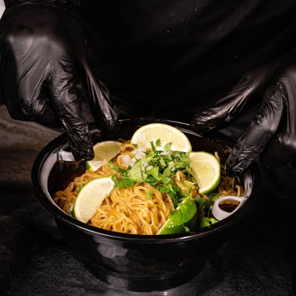 Gloved hands present a bowl of Big Papi's Mexican noodles, garnished with lime, cilantro, and onions.